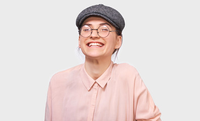 Candid shot of happy young woman, smiling broadly, wears pink shirt and round transparent spectacles. Pretty girl student feels joyful and posing over white studio wall. People and emotions concept