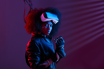 Stylish curly dark haired girl dressed in black leather jacket and gloves is wearing the virtual reality glasses on her head in the dark studio with neon light
