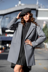 Stylish brunette girl dressed in a fashionable gray dress, a gray jacket and a cap poses in the city street on the sunny day