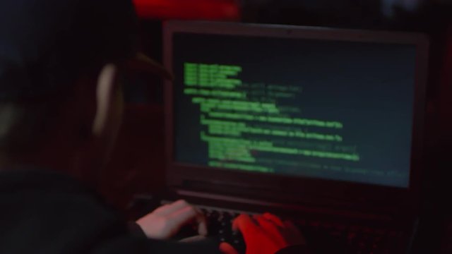 Back view of hacker sitting at laptop, typing on keyboard and breaking into system