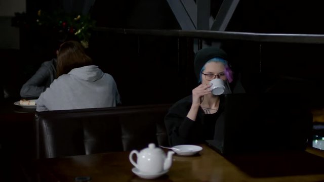 Sequence of videos with young Caucasian woman in hoodie and hat sitting at caf&eacute; table at laptop, typing computer code and drinking tea