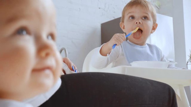 selective focus of kid pointing with finger at brother while boy eating puree in highchair and mother wiping his mouth with napkin