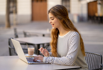 Attractive young woman studying or working in a coffee shop outside with laptop in european city