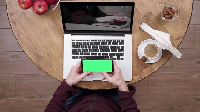 Man in coffee shop watches a video and holds the smartphone with green screen on. Insert any image on the smartphone screen.