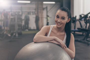 Young happy beautiful woman smiling to the camera, resting at the gym after working out with fitness ball. Cheerful healthy sportswoman relaxing at sports studio after exercising, copy space