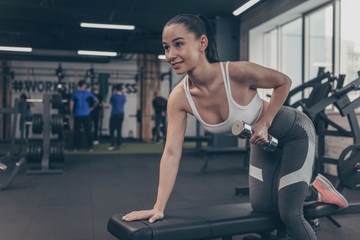 Fototapeta na wymiar Happy beautiful sportswoman smiling joyfully, while exercising with dumbbells at the gym. Stunning cheerful athletic girl lifting weights, copy space. Sporty female enjoying working out with weights