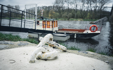 Fototapeta premium Iron old anchor with rope by the river. Ship's knot. In the background a small blue pier and an old shipping boat
