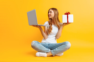 Portrait of an excited girl sitting on the floor with a laptop and a gift box in her hands, on a...