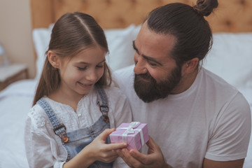 Bearded long haired happy man smiling, giving small gift to his cute little daughter. Adorable little girl receiving a present from her loving father. Celebrating, birthday concept