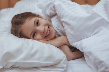 Obraz na płótnie Canvas Lovely little happy girl laughing excitedly to the camera, lying in her bed in the morning. Adorable little girl waking up, smiling to the camera. Children, family, happiness concept