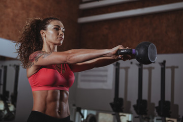 Fototapeta na wymiar Attractive female crossfit athlete working out with kettlebell at the gym. Beautiful sportswoman with perfect fit body lifting kettlebell at crossfit box gym