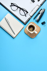 Flat lay, top view office table desk. Workspace with blank clip board, notepad, eyeglasses, office supplies, stapler, pen and coffee cup on blue background. Blog concept