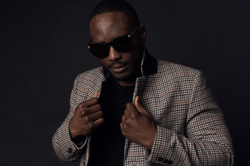 Serious cool man in dark sunglasses, black sweater and brown plaid coat and jeans. Studio, dark key
