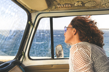 Side view of happy pretty tourist in sunglasses sitting inside a recreational vehicle and looking out of the window. Concept of freedom and relax. Hippie smiling middle age woman in van at seaside
