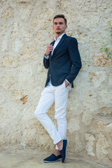 Obraz premium Young handsome man in a suit and white trousers at standing near a stone wall.