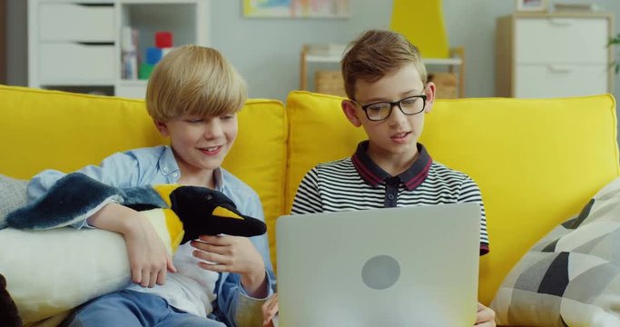 Caucasian teenage small boy in glasses watching something or playing on the laptop and showing this to his friend who hugging pinguin toy on the yellow couch at home.