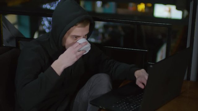 Lockdown of young hooded man sitting in bar at computer, typing on keyboard and drinking hot tea