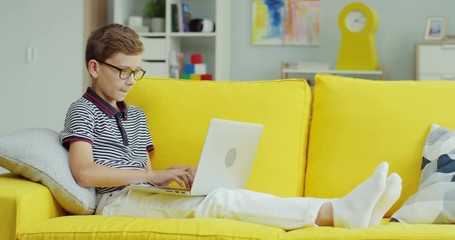 Caucasian teen boy in glasses sitting on the yellow sofa at home and playing on the laptop computer, then winning.
