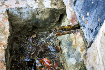 close up of a fountain where across a half pipe flows fresh spring water from a natural source. The water jet falls in a small black hole at the bottom of the fountain with many dry leaves, horizontal