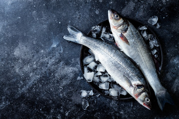 Fresh raw seabass fish on black stone background with ice. Culinary seafood background. Top view,...