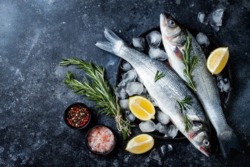 Fresh raw seabass fish on black stone background with spices, herbs, lemon and salt. Culinary...