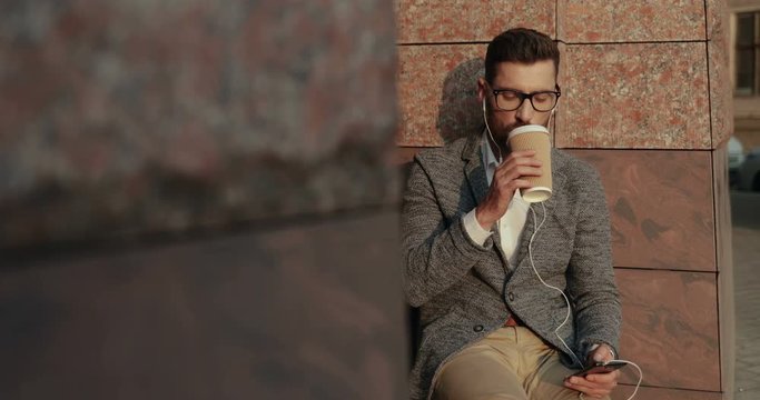 Handsome young Caucasian businessman in glasses sitting at the building on the steps with a coffee and listening to the music on the smartphone via headphones.