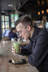 Young stylish man enjoys a cake and a drink while sitting in a cafe with large windows