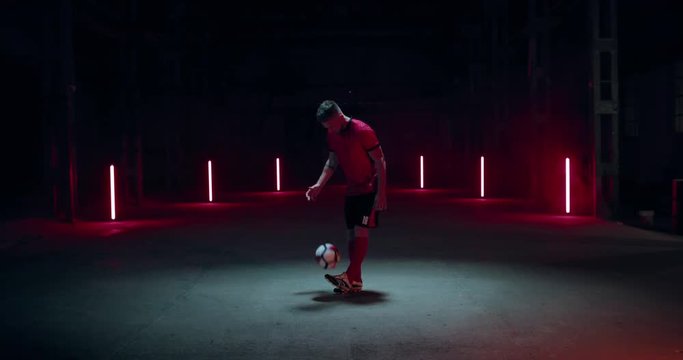 WIDE FIXED Caucasian professional football soccer player posing against dark background in a large abandoned warehouse. 4K UHD 60 FPS SLOW MOTION