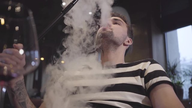 Handsome man enjoying hookah sitting in modern restaurant cafe bar. Confident client exhales clouds of steam sitting in a hookah close up. Handsome man relaxing alone