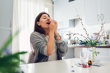 Spring allergy. Young woman sneezing because of flowers surrounded with pills on kitchen. Seasonal allergy concept.
