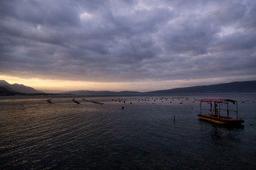 Shellfish breeding on Mediterranean sea. Mussel farm with boat on Adriatic coast. Oyster beds at low tide in oyster farm in early morning golden light. Dramatic sky with grey clouds over calm water.