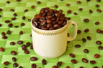 White cup for coffee full of coffee beans on green plastic tablecloth.