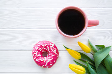 Tasty pink doughnut and black coffee in living coral color cup in white background tabe, free space for text. Top view. Bouquet of yellow tulips decorations. 