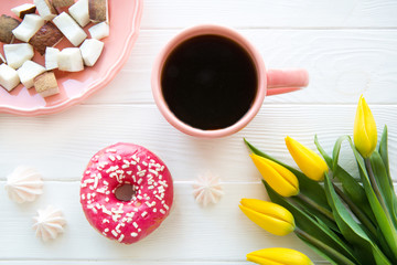 A pink strawberry doughnut, coconut pieces and meringue on white wooden table, top view. Yellow flower tulips decorations. 