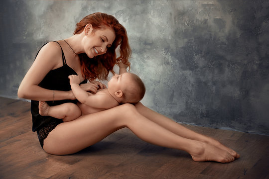 Beautiful redhead girl, mother, in black underwear sits on the floor and plays with a naked baby, smiling