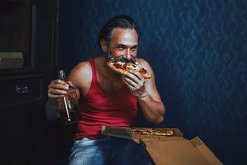 Selbstklebende Fototapeten Brutal Bearded Man at Home Eating Pizza and Drinking Alcohol. © Svyatoslav Lypynskyy