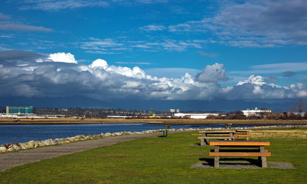 Picnic area with dining tables an the River Bank of Fraser River, Vancouver Airport on the opposite bank of the river, residential areas of Vancouver City on the horizon against a cloudy sky