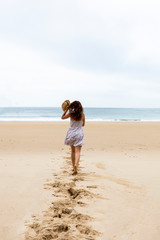 Fototapeta na wymiar Girl in hat and white dress running towards water on the empty beach