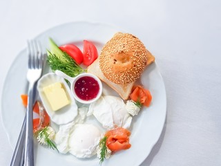 Breakfast on a white plate. Poached eggs, lightly-salted salmon, cheese cream, butter, slices of tomato and cucumber , dill, bun with sesame and toast.