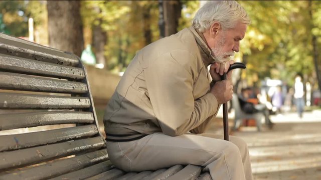 Portrait of sad tired senior man sitting on bench in autumn park leaning on cane