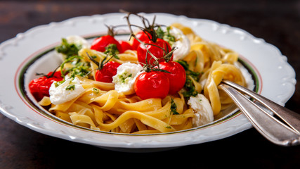 Fettuccine pasta with cherry tomatoes, mozzarella and pesto sauce. Hot dish with baked vegetables on a dark background.