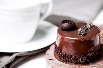 A delicious chocolate cake with chocolate pieces lies on a wooden stand next to a white cup, which stands on a white table
