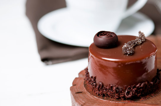 A delicious chocolate cake with chocolate pieces lies on a wooden stand next to a white cup, which stands on a white table