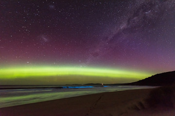 An incredible display of the Aurora Australis or Southern Lights over a beach in Tasmania with bioluminescence turning the breaking waves bright blue.