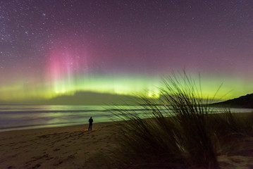 One person in silhouette standing in awe of an incredible display of the Aurora Australis or Southern Lights, with bioluminescence turning the breaking waves bright blue.