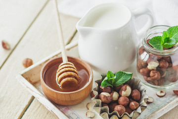 Honey in the wooden bowl, mint leaves, hazelnuts and jar with milk on the wooden tray