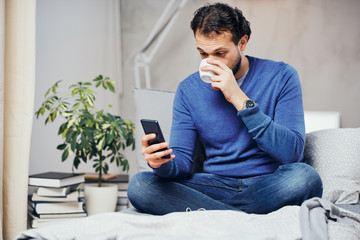 Attractive smiling arab man dressed casual sitting on bed in bedroom, drinking coffee and using smart phone for texting.