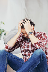 Attractive arab man in jeans and plaid shirt sitting on the floor in bedroom and holding head. Men's issues concept.