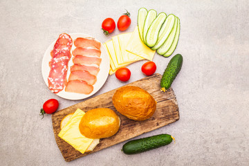 Homemade sandwich in bun, step by step preparation. Salami, dried pork, tomato, cucumber, cheese ingredients. On wooden board and stone background, top view.