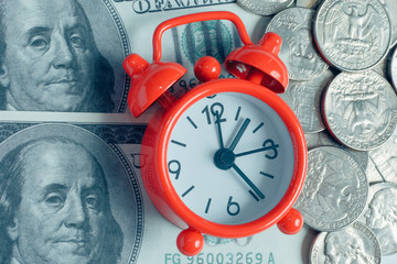 Concept of saving money in business. Red vintage clock on hundred dollar bills and coins. Toned.